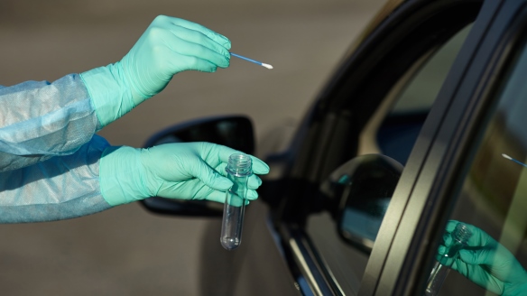 Photo of hands wearing gloves holding a swab at the open window of a car to test the driver for COVID-19
